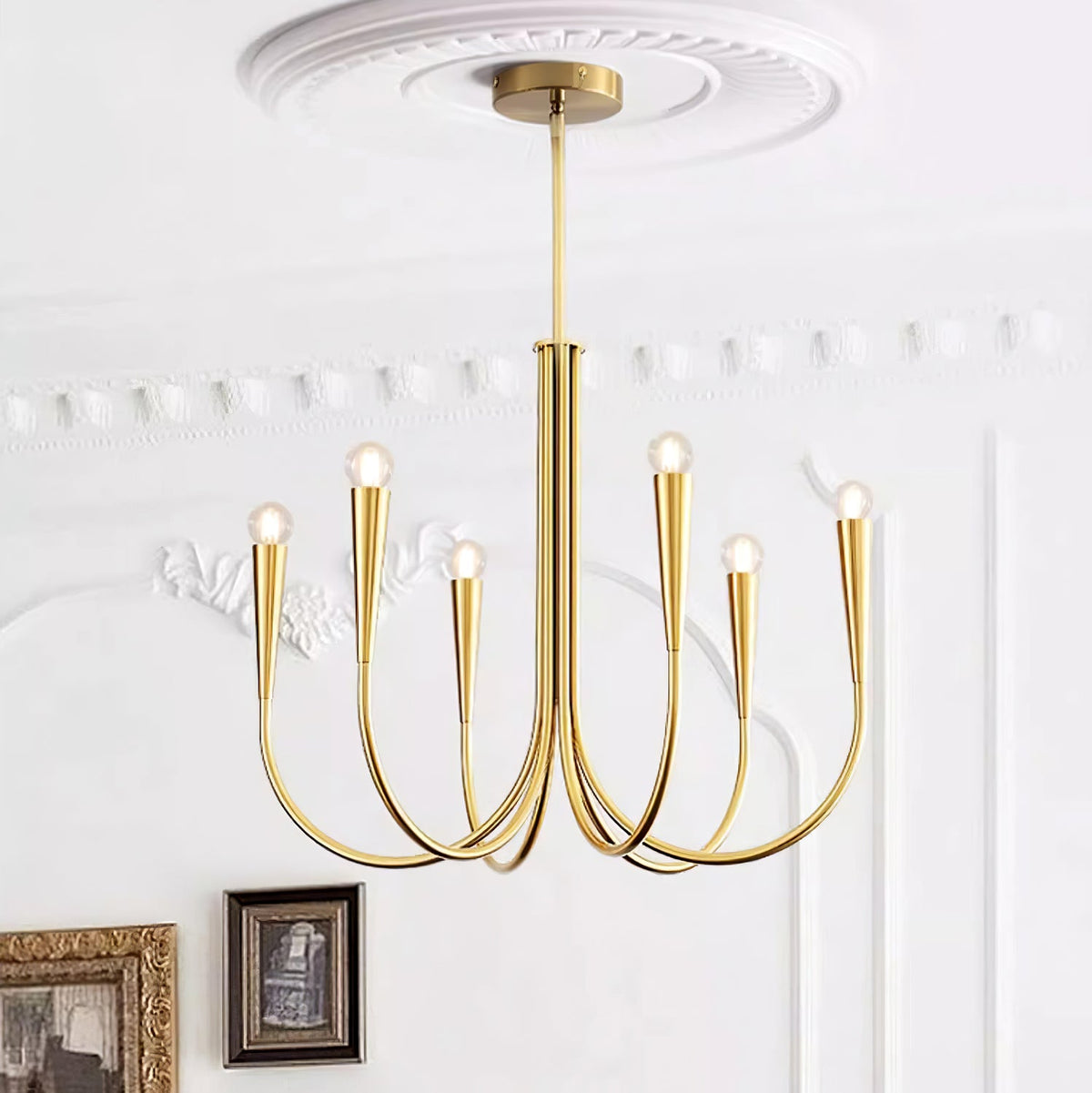 Looking up at a Clérachic chandelier in a brass tone, with a plaster ceiling rose, white wall moulding and framed pictures below.