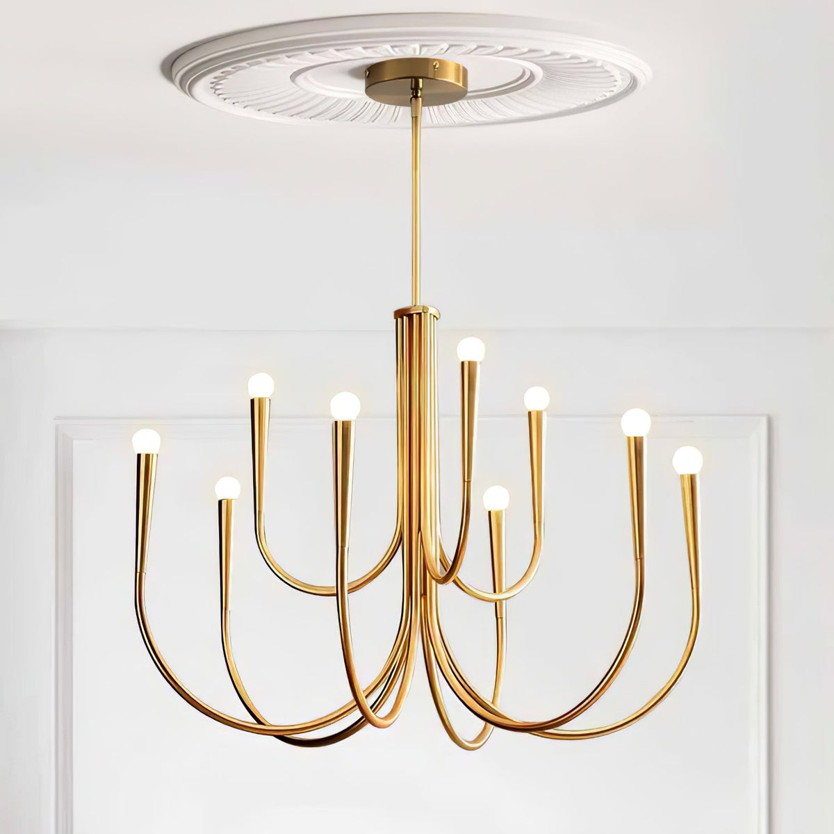 A Clérachic chandelier with a brass-toned finish, hung from a plaster ceiling rose against white panelled walls.