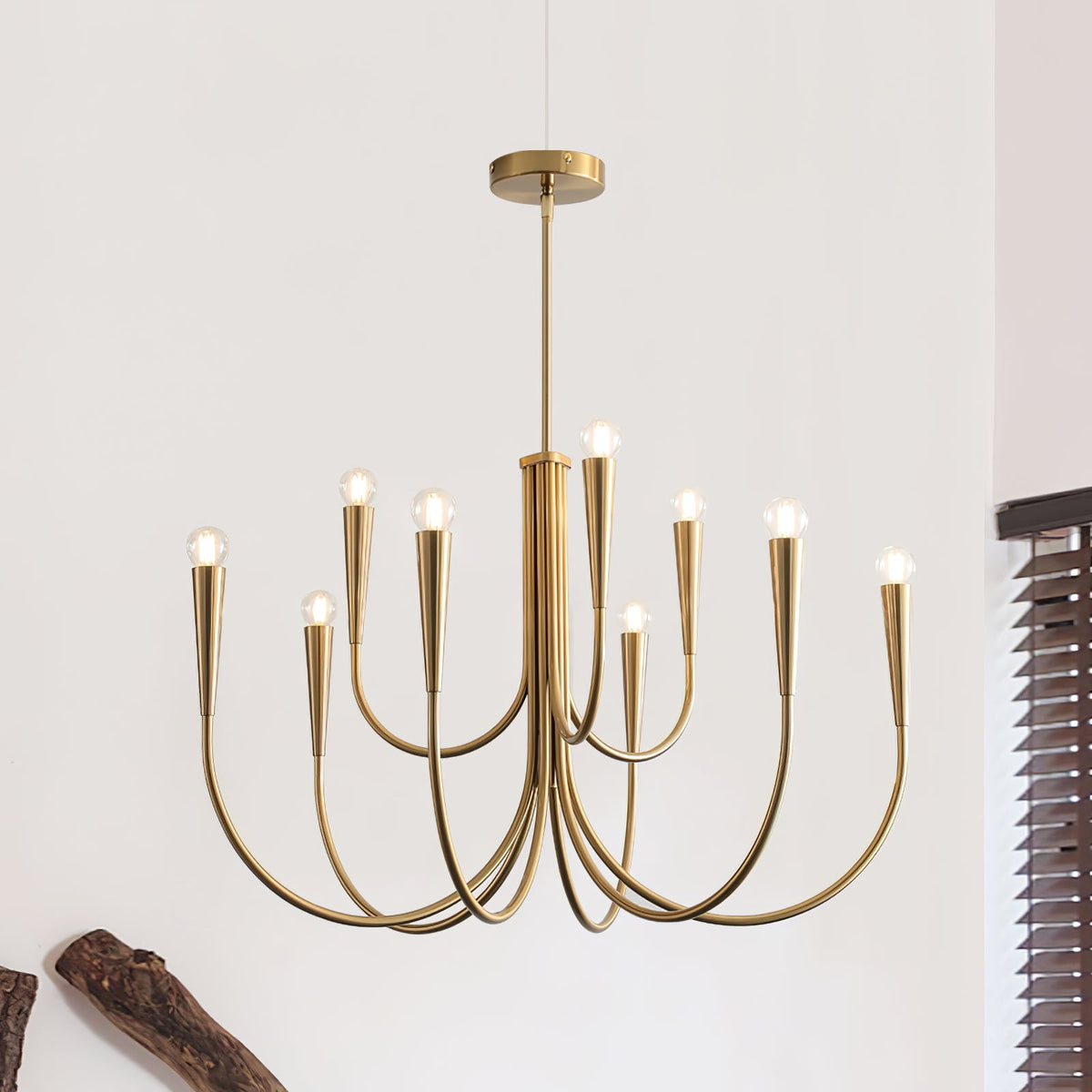 Brass-toned Clérachic chandelier on a white wall, with a dark wooden log at the lower left and a blind at the right edge.