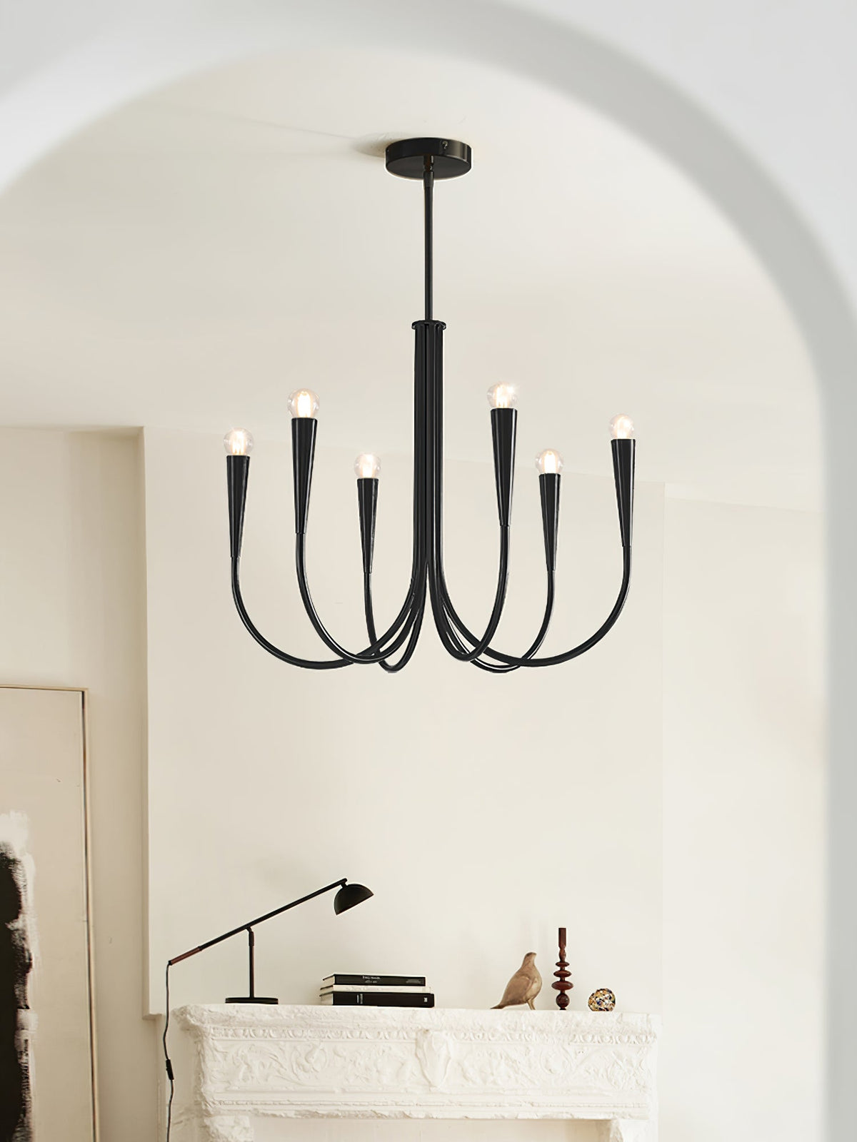 A black Clérachic chandelier in an arched white room, above a mantel with a desk lamp and small ornaments.