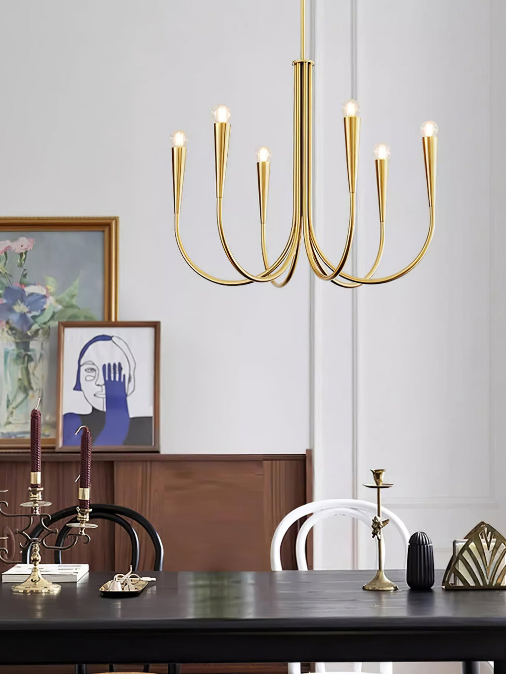 A smaller brass-toned Clérachic chandelier above a black table with candlesticks, a framed print and a wooden sideboard.