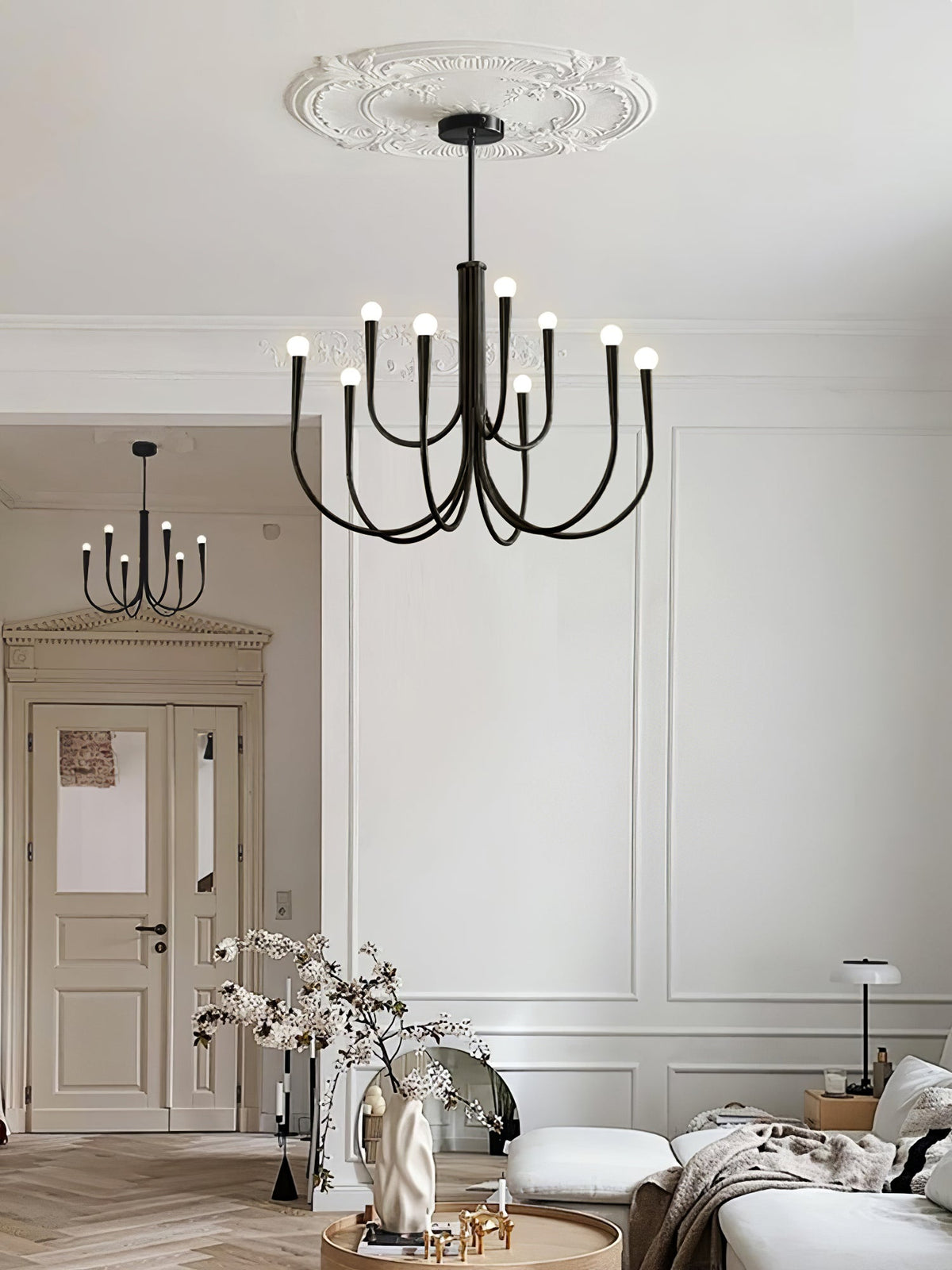 Black Clérachic chandelier under a plaster ceiling rose in a white room, with a sofa and a vase in the room behind.