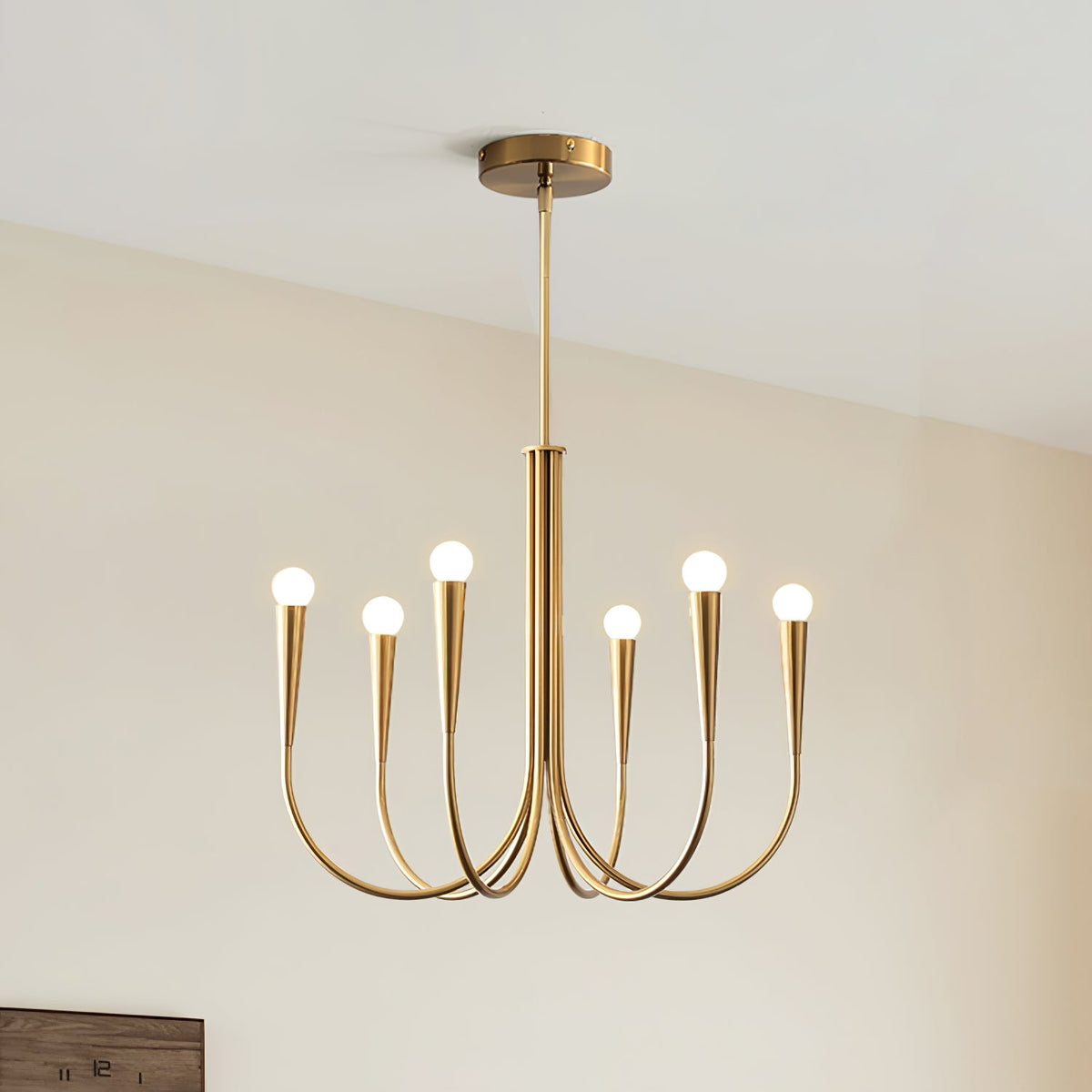 Brass-toned Clérachic chandelier with six arms and a rod, hung against a cream wall with the corner of a wooden frame below.