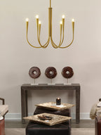Small brass-toned Clérachic chandelier with six arms, seen against a warm wall with a console table and ornaments below.