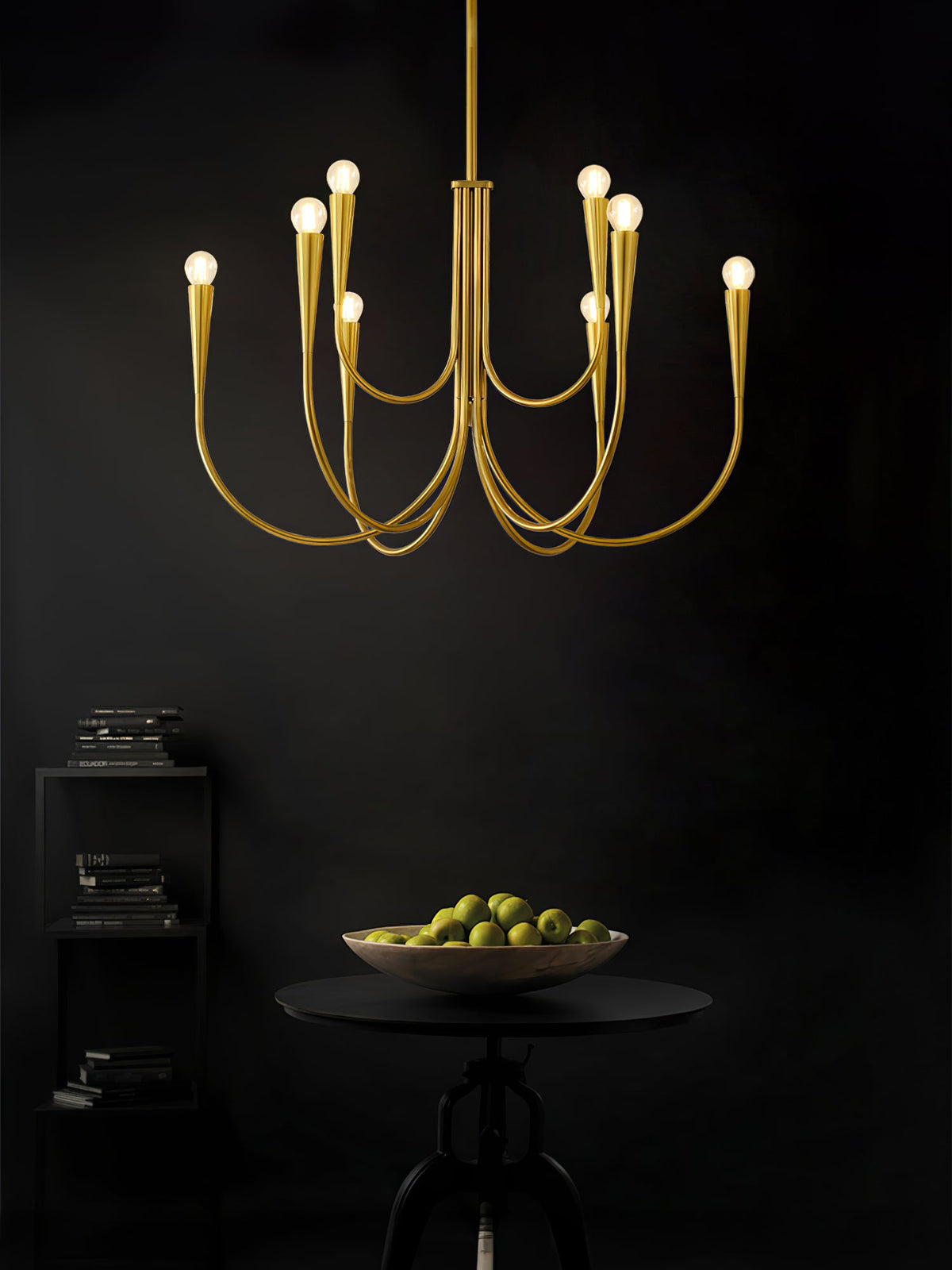 Brass-toned Clérachic chandelier lit against a very dark room, above a round black table with a bowl of limes.