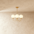 Clérachic three-light chandelier with white glass globes on curved brass-toned arms, hung from a rod against a limewash wall.