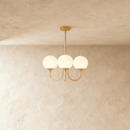 Clérachic three-light chandelier with white glass globes on curved brass-toned arms, hung from a rod against a limewash wall.