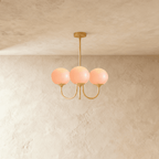 Clérachic three-light chandelier with pale pink glass globes lit warm on curved brass-toned arms and a slim ceiling rod.