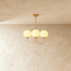 Clérachic three-light chandelier glowing with cream-toned glass globes on curved brass-toned arms against a limewash backdrop.