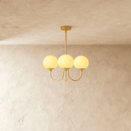 Clérachic three-light chandelier with softly lit butter-yellow glass globes on curved brass-toned arms and a slim stem.