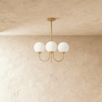 Clérachic three-light chandelier with unlit white glass globes on curved brass-toned arms, shown against a limewash wall.