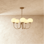 Clérachic five-light chandelier with softly glowing cream oval glass shades on sweeping bronze-toned arms, limewash wall behind.