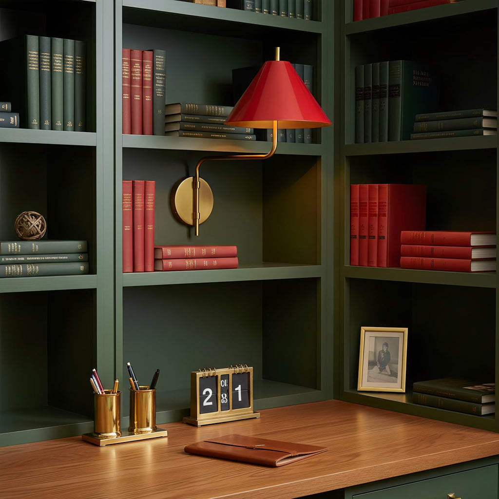 Red-shaded Clérachic wall light on a dark green bookcase over a wooden desk, with red-spined books and brass desk pots below.