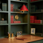 Red-shaded Clérachic wall light on a dark green bookcase over a wooden desk, with red-spined books and brass desk pots below.