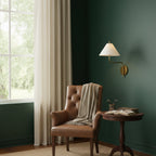 White-shaded Clérachic wall light on a deep green wall above a small round table and a tan leather armchair by a window.