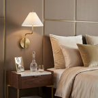 White-shaded Clérachic wall light beside a padded taupe headboard with brass trim, above a marble-topped bedside table with a decanter.