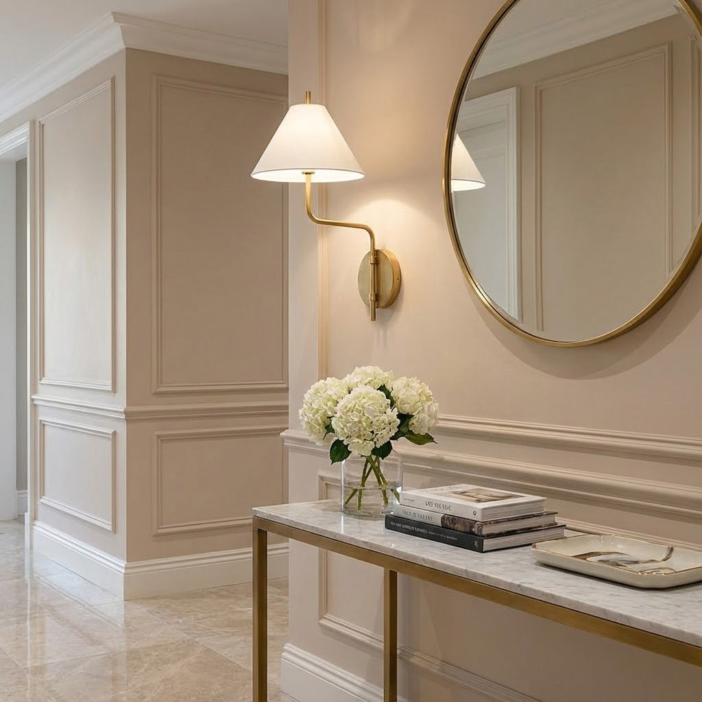 White-shaded Clérachic wall light in a panelled hallway, beside a round brass-framed mirror and over a marble console with white hydrangeas.