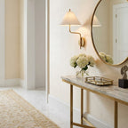White-shaded Clérachic wall light by a tall round mirror in a bright hallway, above a marble console with white hydrangeas and a brass tray.