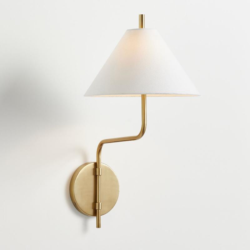 Clérachic wall light with a white fabric shade, a bent gold arm and a round gold backplate, on a plain white wall.
