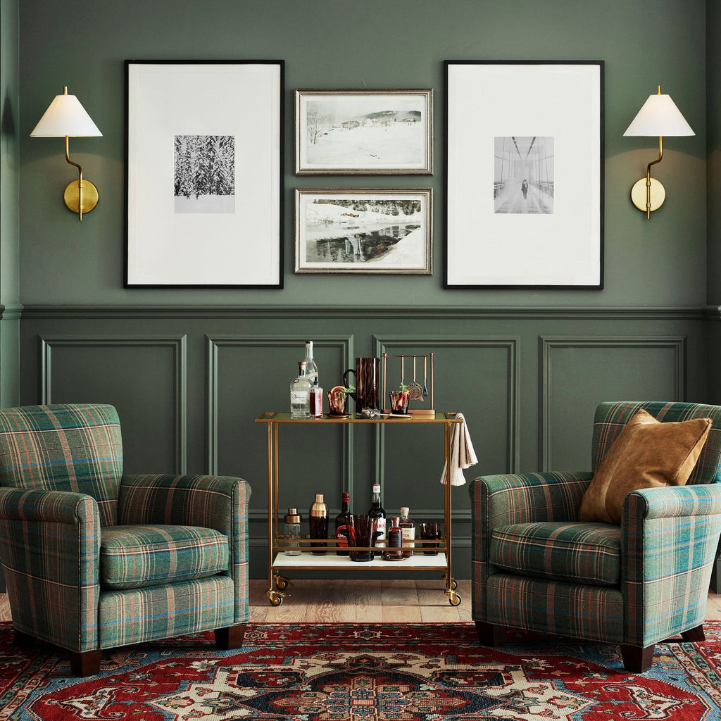 Two white-shaded Clérachic wall lights either side of framed prints on a green panelled wall, above a bar cart and plaid armchairs.