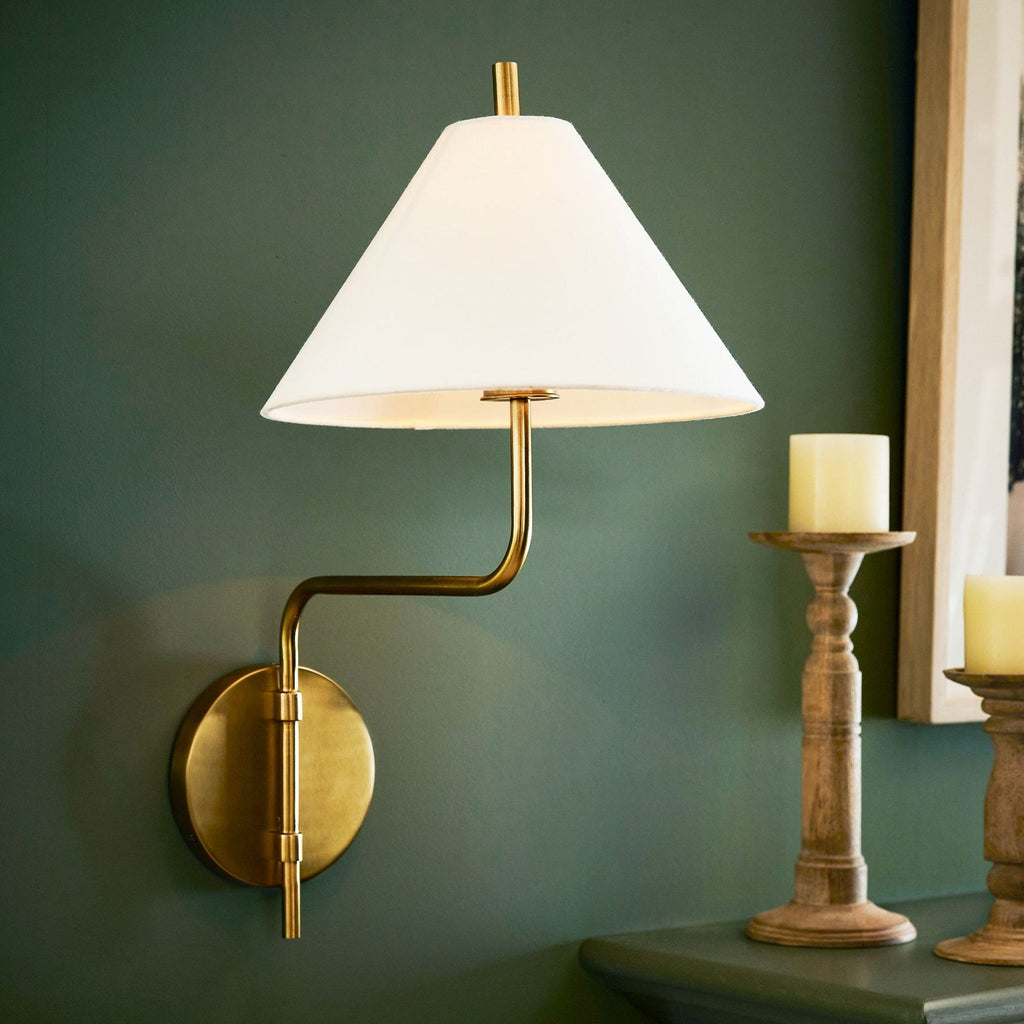White-shaded Clérachic wall light on a deep green wall beside a candle holder on a console, its gold arm bending out from a round plate.