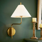 White-shaded Clérachic wall light on a deep green wall beside a candle holder on a console, its gold arm bending out from a round plate.