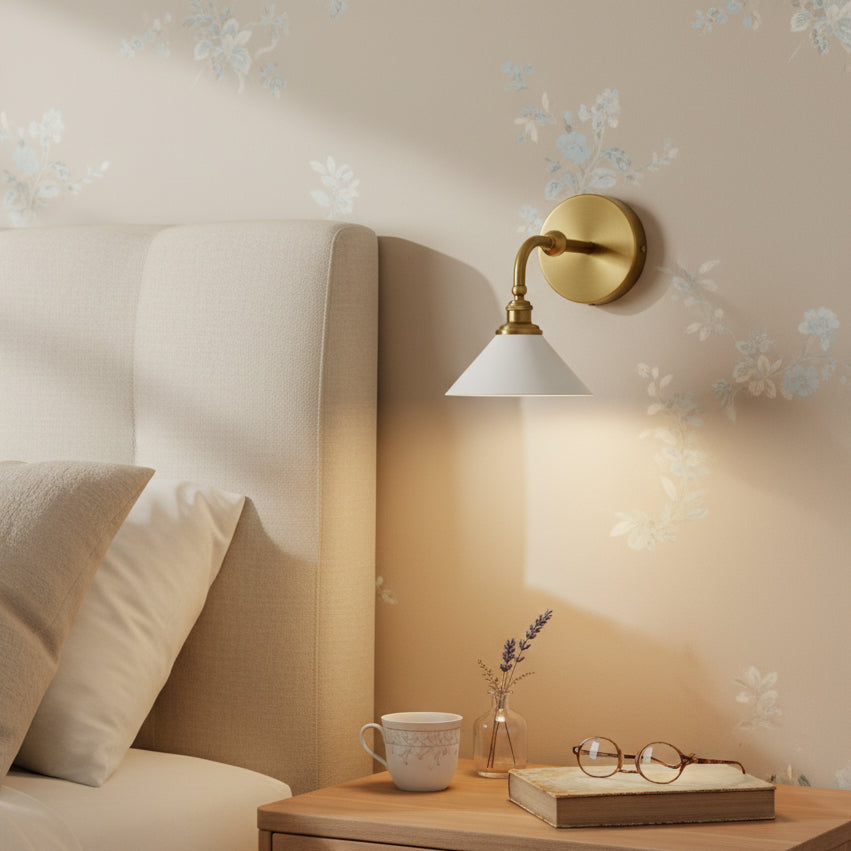 This Clérachic wall light, with a brass curved arm and a white cone shade, glows beside a cream headboard and a floral wallpaper.