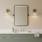 A pair of Clérachic wall lights with brass arms and white cone shades flank a black-framed mirror above a sage-green vanity.