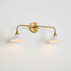 Brass double-arm Clérachic wall light with two white conical shades, both lit, on a plain pale wall.