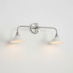 Nickel double-arm Clérachic wall light with two white conical shades, lit, on a plain pale wall.