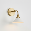 Close view of the brass round backplate, curved arm and white cone shade of this Clérachic wall light, lit on a pale wall.