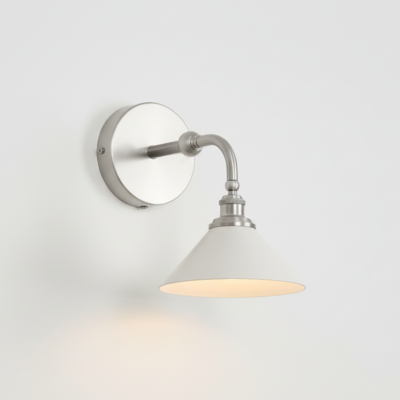Nickel version of this Clérachic wall light, with a round nickel backplate, a curved arm and a white cone shade, lit on a pale wall.