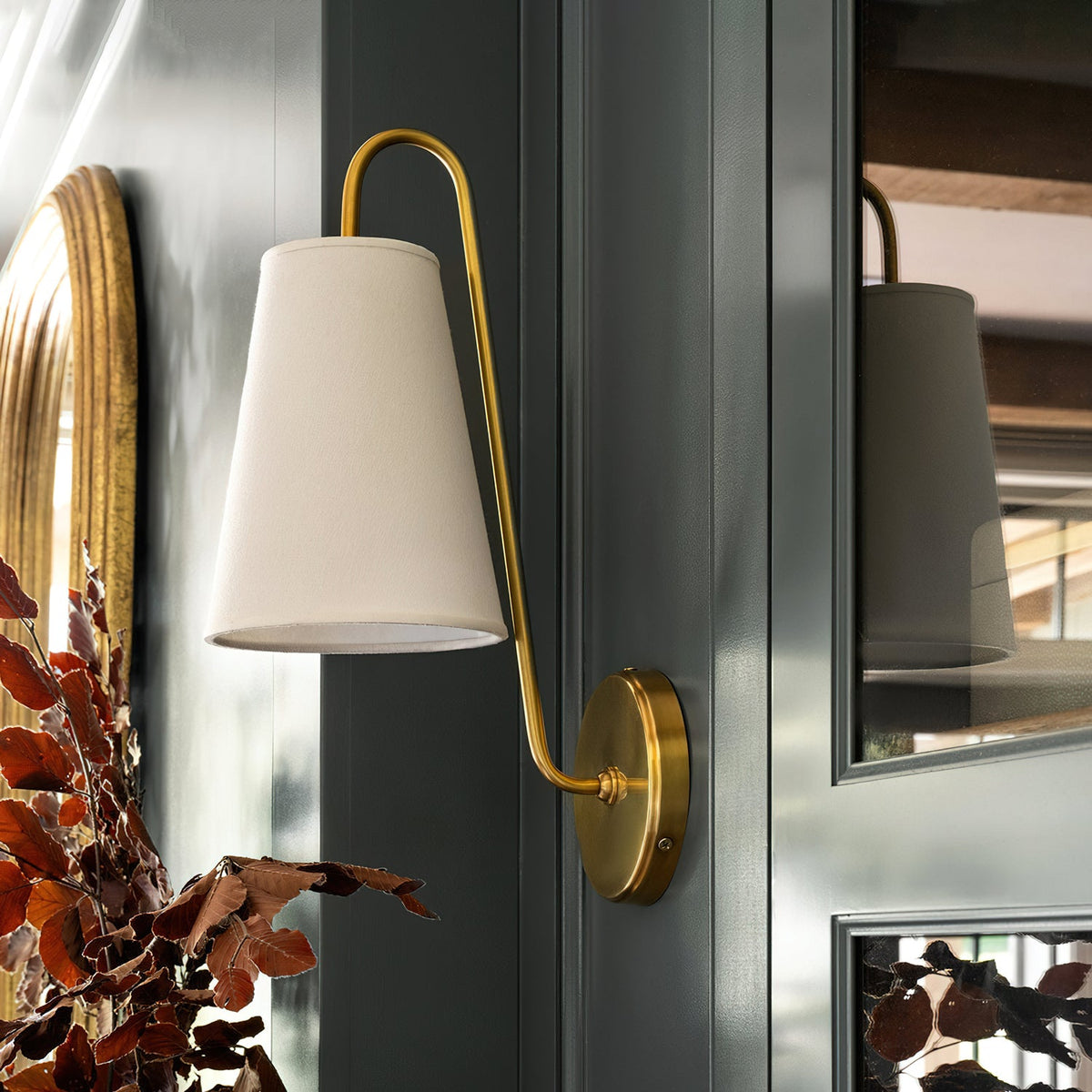 Close view of a brass Clérachic wall light with a cream tapered shade, beside a gilt-framed mirror and a dark green door.