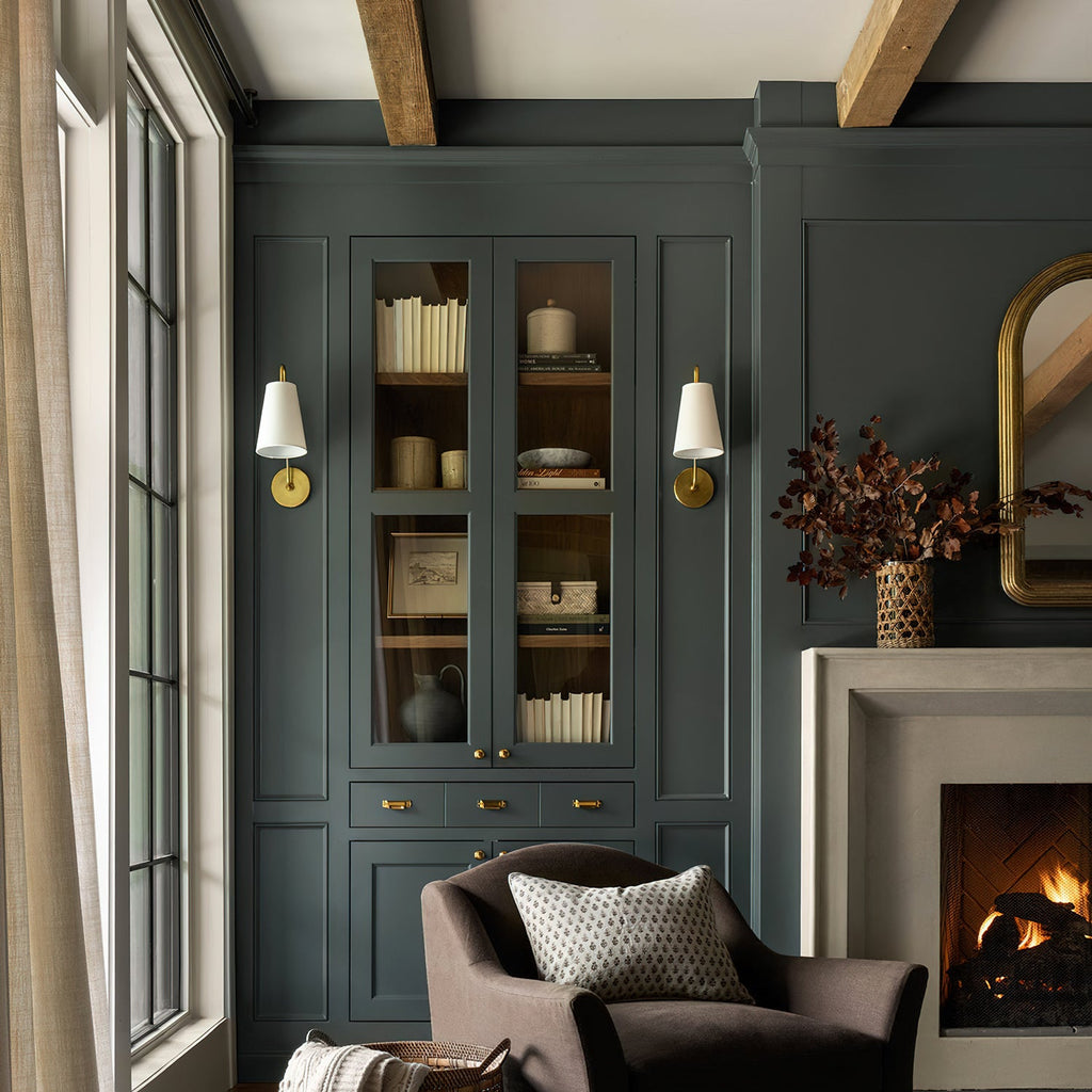 Two brass Clérachic wall lights with white shades, either side of a green built-in cabinet, beside a stone fireplace.