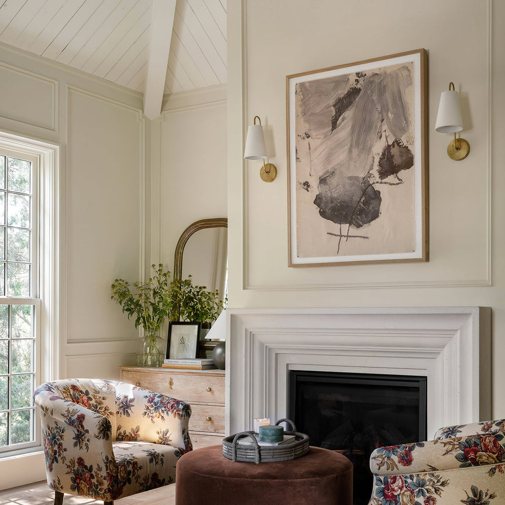 A pair of brass Clérachic wall lights with white tapered shades on a cream wall, above a fireplace with floral armchairs.