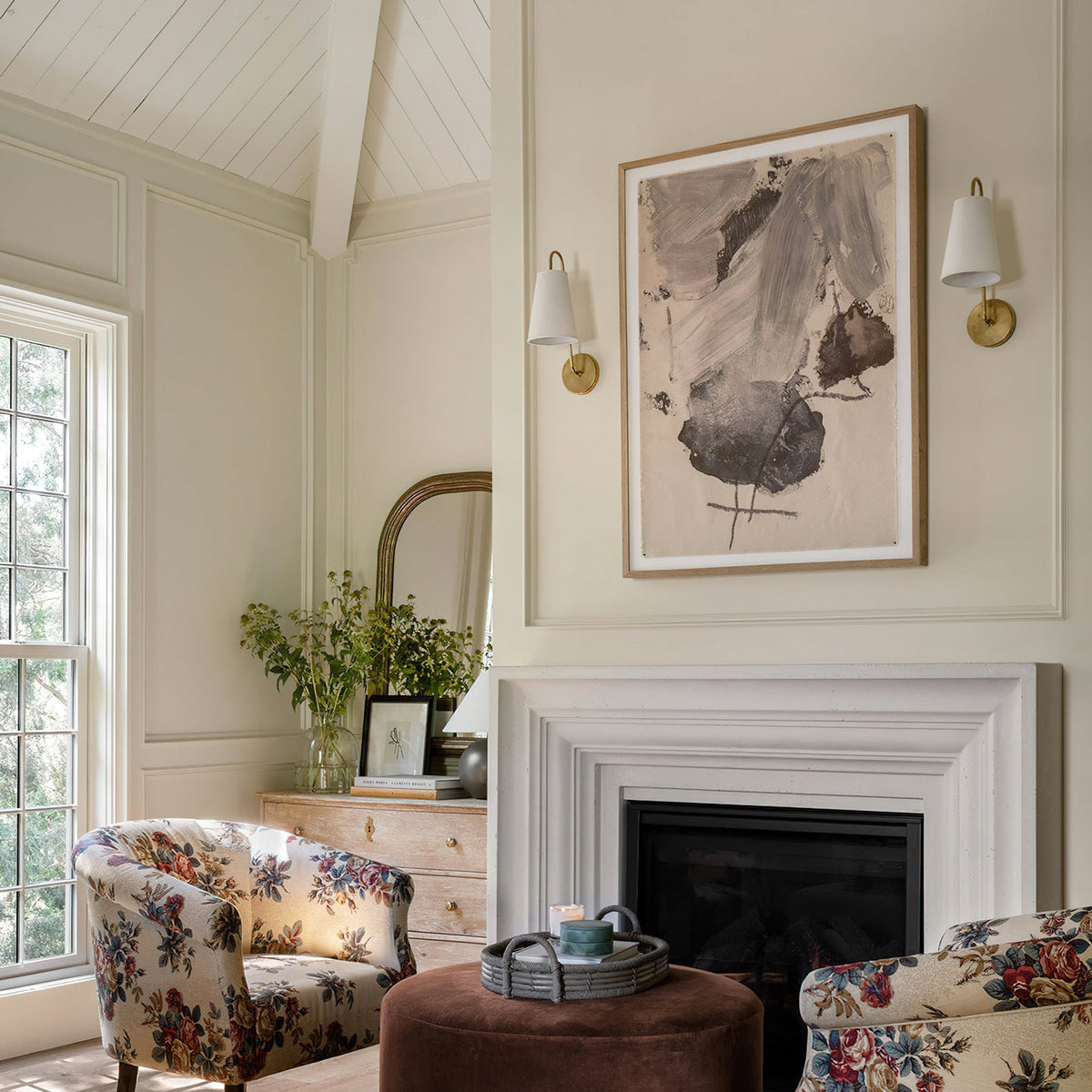 A pair of brass Clérachic wall lights with white tapered shades on a cream wall, above a fireplace with floral armchairs.