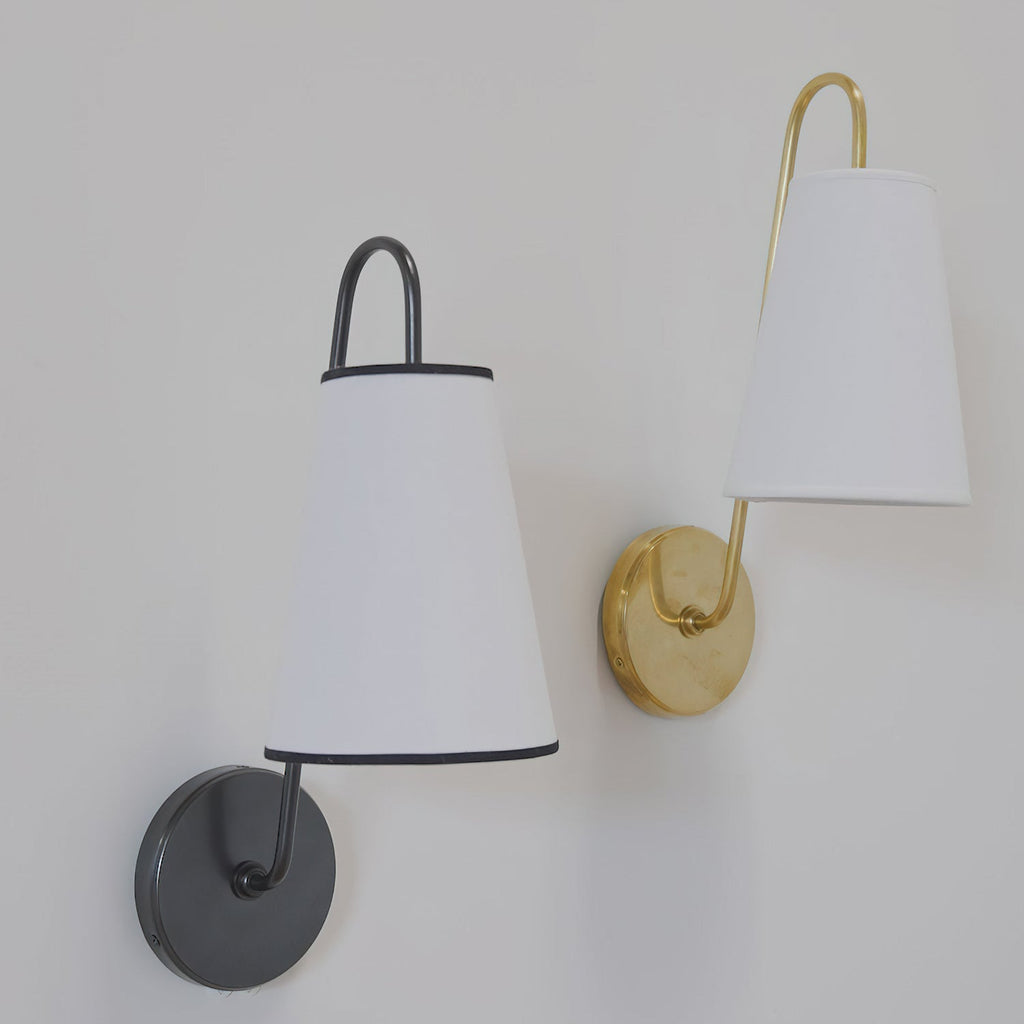 A black and white Clérachic wall light beside a brass and white one, each with a tapered fabric shade, on a pale grey wall.