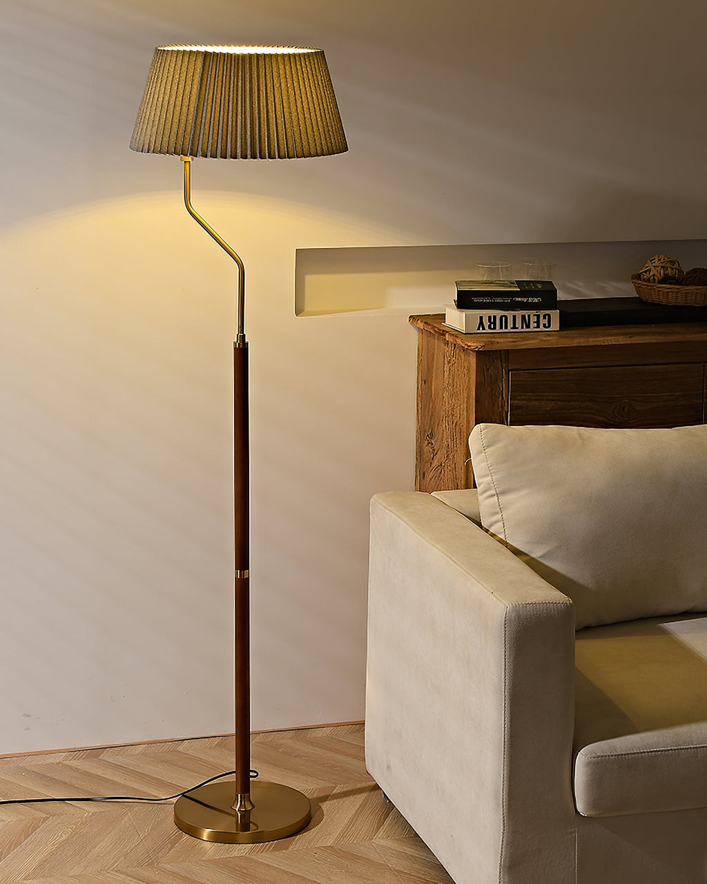 A Clérachic floor lamp with a pleated shade lit warmly, its brass base on herringbone flooring beside a linen armchair.