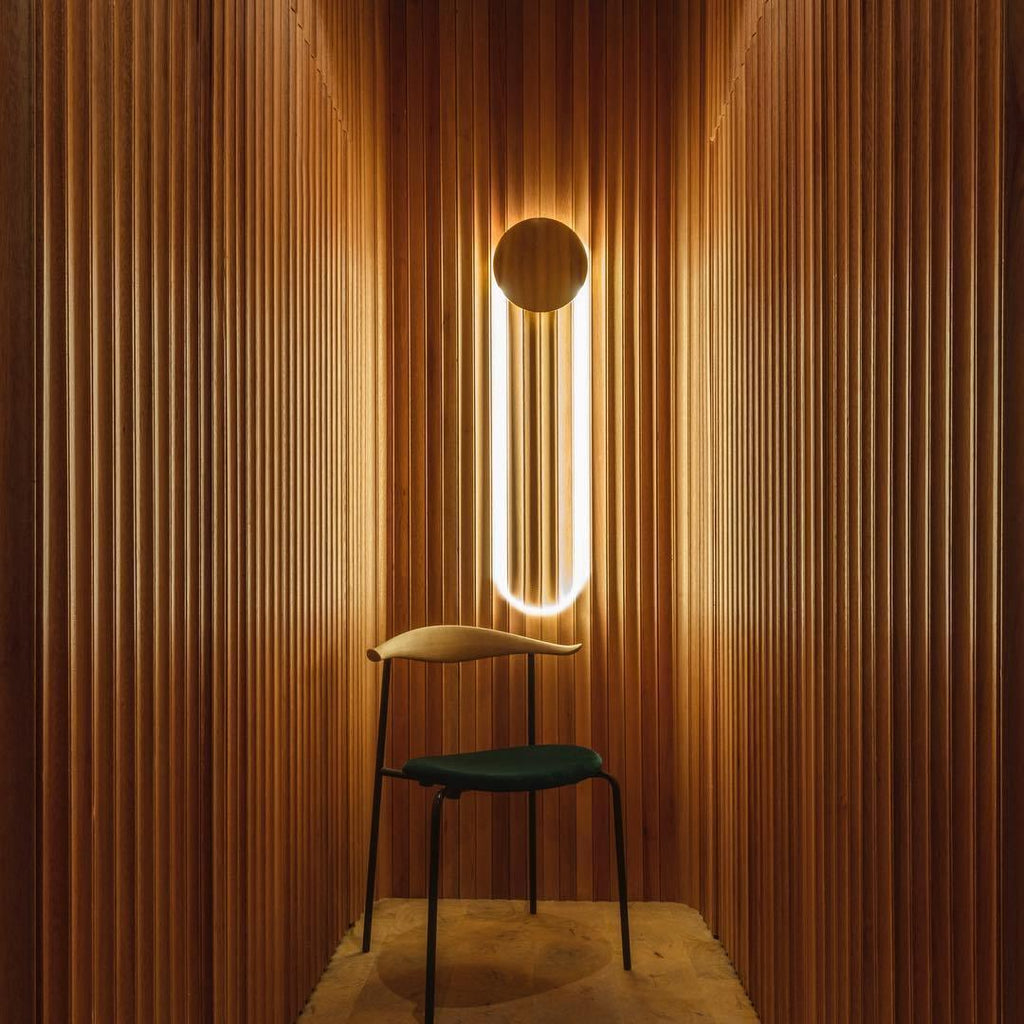 Between fluted timber panels, a Clérachic wall light glows warm in a narrow space behind a dark green chair with a slim wooden frame.