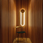 Between fluted timber panels, a Clérachic wall light glows warm in a narrow space behind a dark green chair with a slim wooden frame.