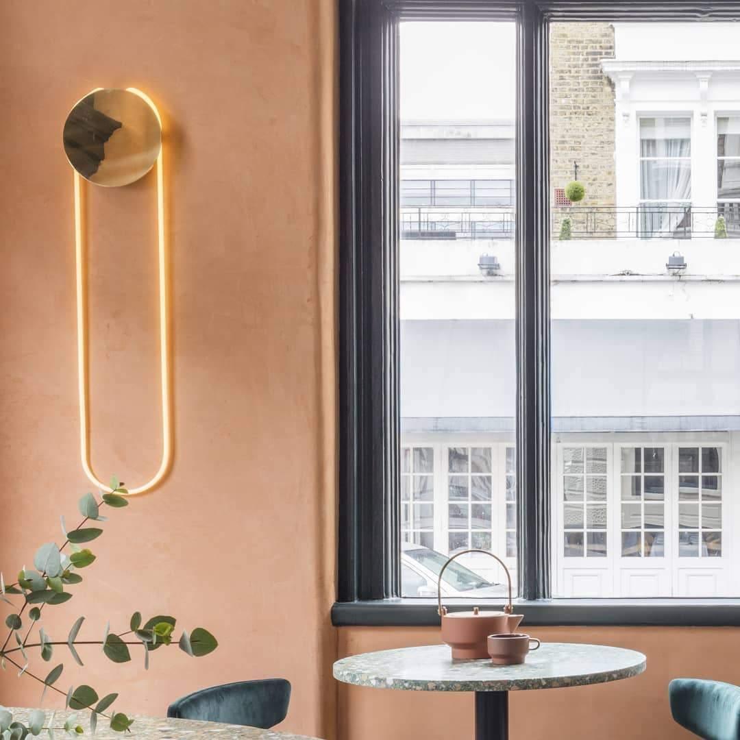 A Clérachic wall light with a gold disc and a warm LED loop on a terracotta wall, beside a tall black-framed window and a small round table.