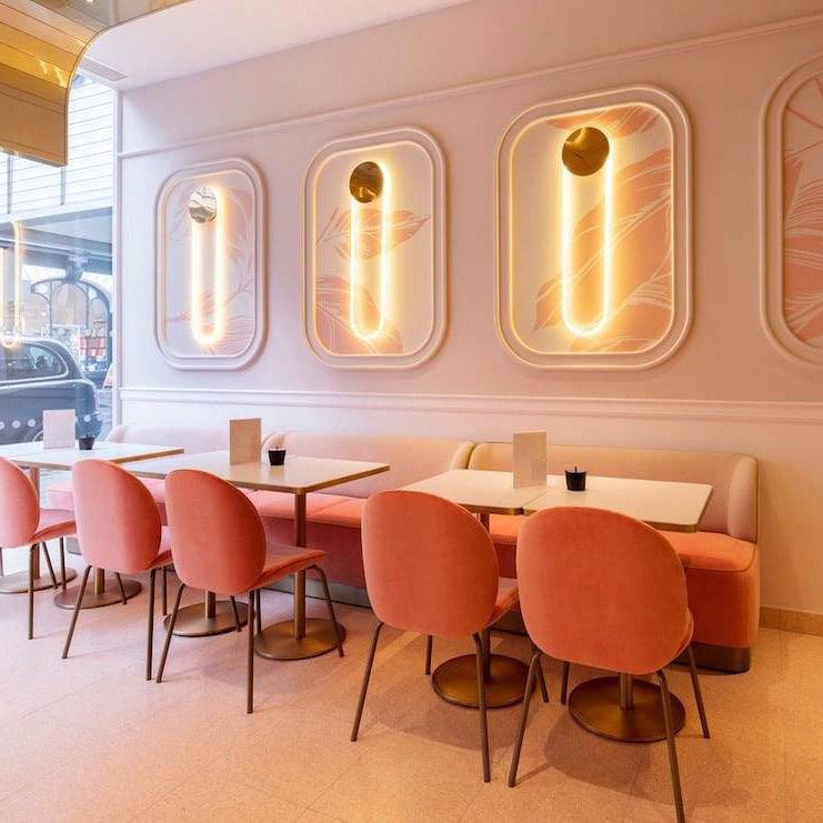 Three Clérachic wall lights glow warm in recessed panels above a row of pink dining chairs and banquette seating in a pale pink café.