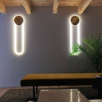 Two Clérachic wall lights with gold discs and bright white LED loops on a grey wall, above a low bench and beside a large palm.