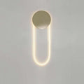 This Clérachic wall light lit warm on a pale grey wall, its brushed gold disc ringed by a faint halo above a long LED loop.