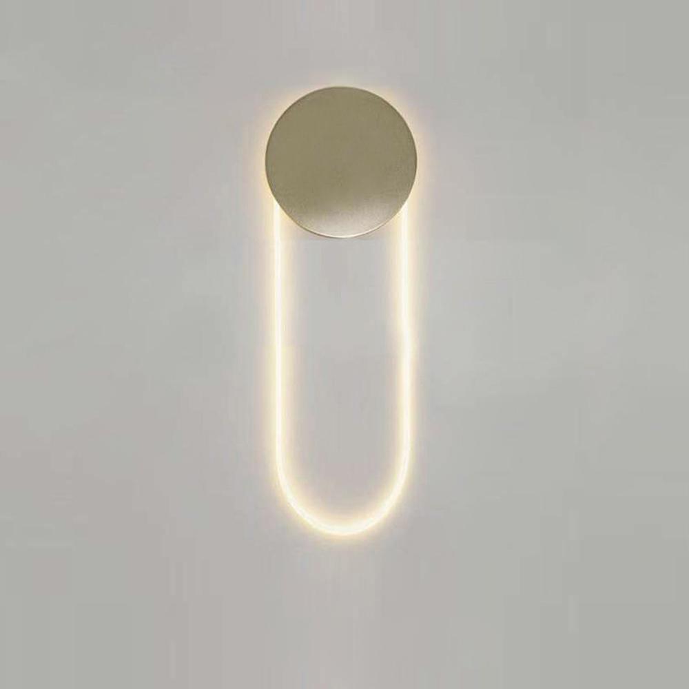 This Clérachic wall light lit warm on a pale grey wall, its brushed gold disc ringed by a faint halo above a long LED loop.