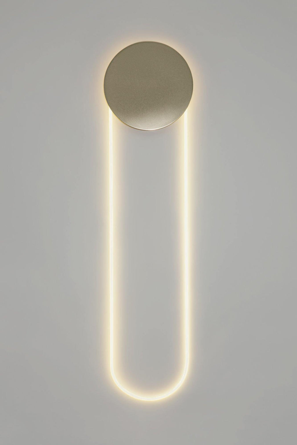 A tall Clérachic LED loop lit warm white under a brushed gold disc, against a light grey wall with a plain white edge.