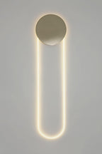 A tall Clérachic LED loop lit warm white under a brushed gold disc, against a light grey wall with a plain white edge.