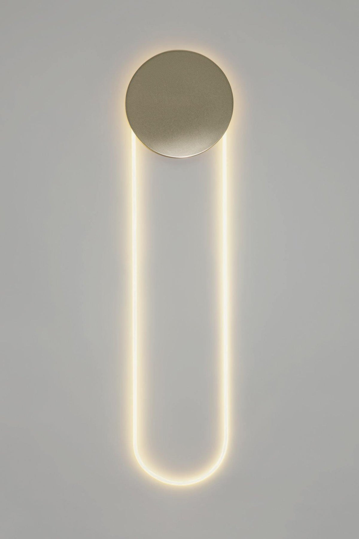 A tall Clérachic LED loop lit warm white under a brushed gold disc, against a light grey wall with a plain white edge.