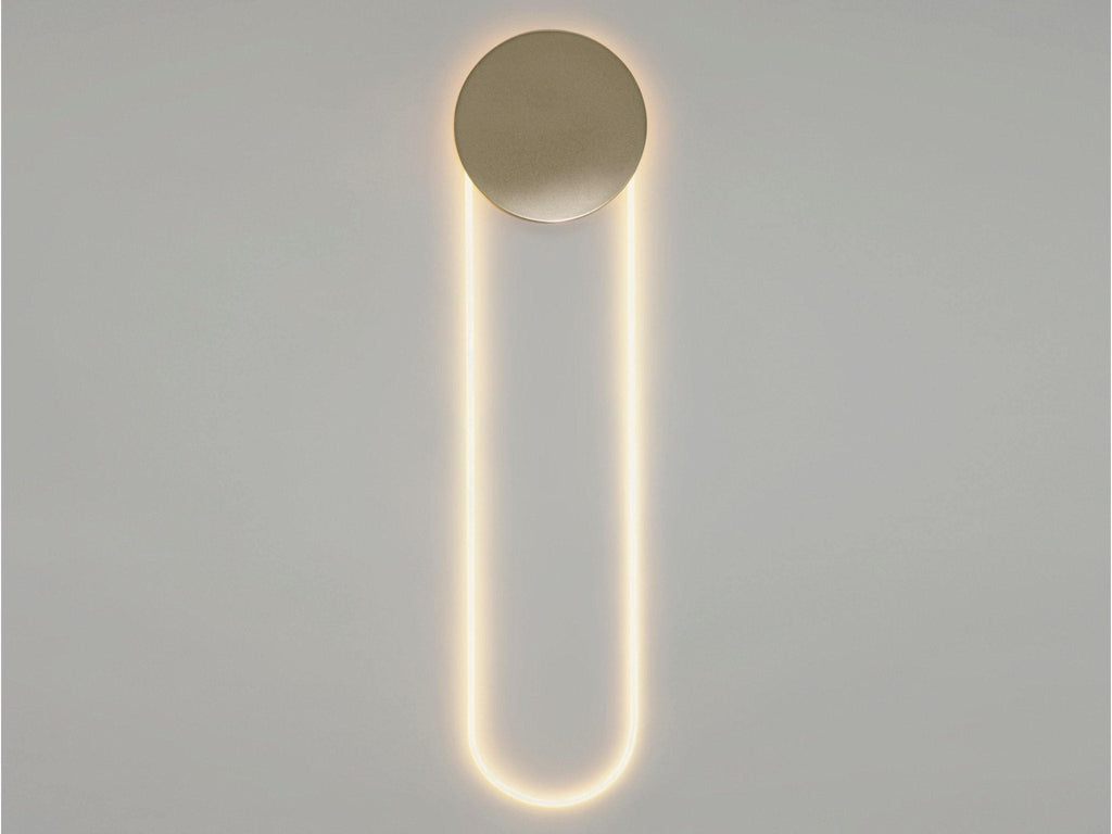 Straight-on view of this Clérachic wall light in a warm glow, a gold disc above a long LED loop on a pale grey wall.