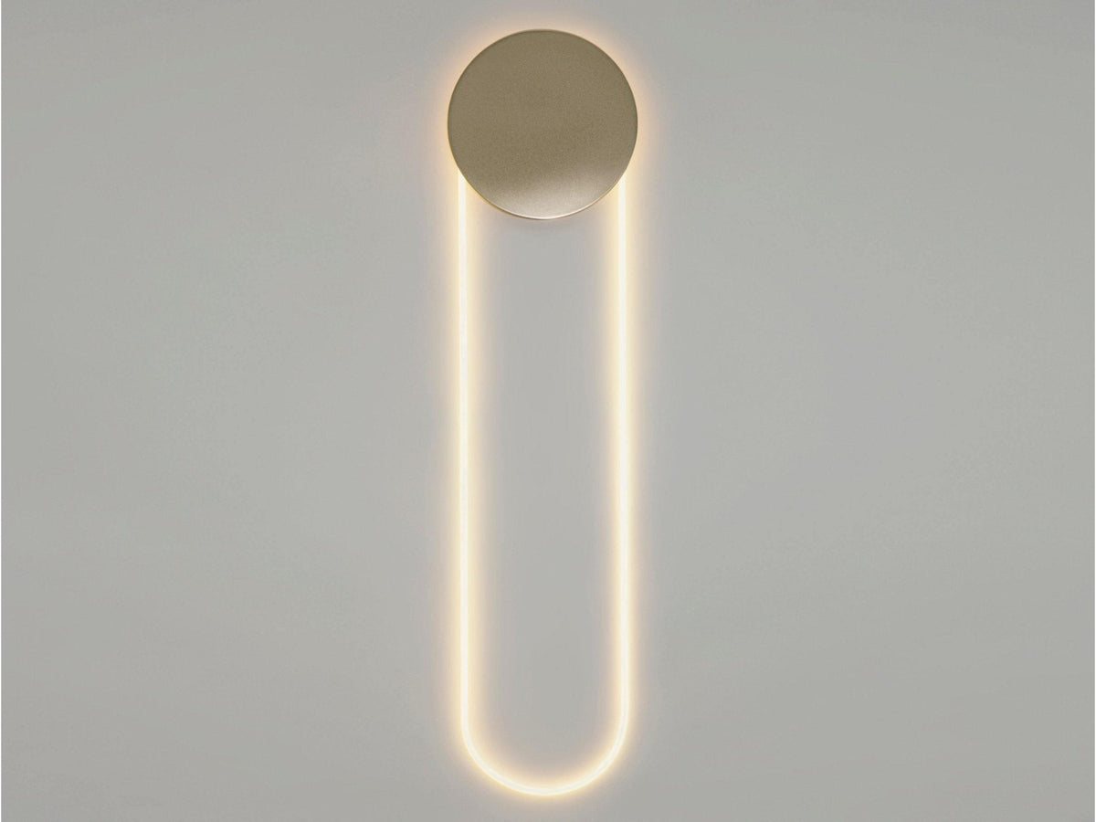Straight-on view of this Clérachic wall light in a warm glow, a gold disc above a long LED loop on a pale grey wall.